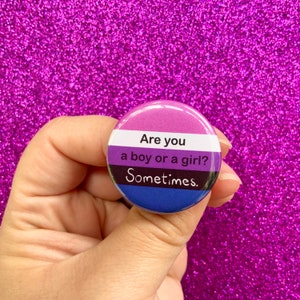 May include: A round button with the colors of the genderqueer flag. The button has the text "Are you a boy or a girl? Sometimes."