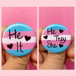 May include: Two round buttons with the transgender pride flag colors. The left button says "He It" with black hearts. The right button says "He They She" with black hearts.