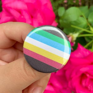 May include: A circular pin-back button featuring a horizontal striped pattern. The stripes are arranged in order from top to bottom as green, light blue, white, yellow, magenta, and dark gray. The surface of the button has a glossy finish.