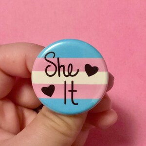 May include: A blue, pink, and white striped button with the text "She It" and two black hearts.