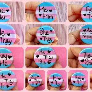 May include: A set of 12 round buttons with the transgender pride flag colors. Each button has a different combination of pronouns written in black text with hearts. The pronouns include: She/Her, He/Him, They/Them, He/They, She/They, Any Pronouns, They/He, He/They/It, She/They/It, It/Its, They/It, He/It.