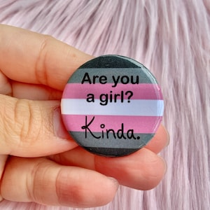May include: A round button with a pink, white, and gray striped design. The text "Are you a girl? Kinda." is written in black on the button.