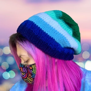 May include: A knitted beanie in shades of blue, green, and white. The hat is worn at an angle, with the top of the hat in green. The person is wearing a face mask with the words "LOVE WINS" in a rainbow of colors.