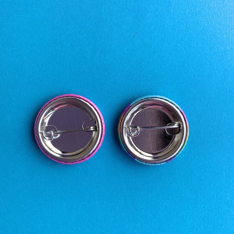 May include: Two round button pins with rainbow colored designs. The back of the pins are silver with a metal pin back.