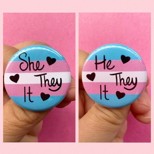May include: Two round buttons with the transgender pride flag colors. The left button says "She They It" and the right button says "He They It".