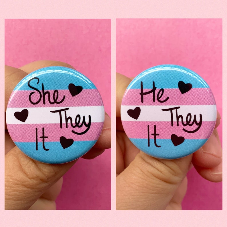 May include: Two round buttons with the transgender pride flag colors. The left button says "She They It" and the right button says "He They It".