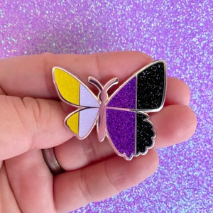 May include: A butterfly-shaped enamel pin featuring a metallic silver outline. The left wing is divided into vertical sections of glittery yellow and light purple, while the right wing features vertical sections of glittery dark purple and black.