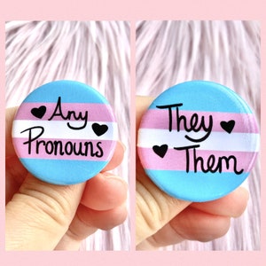 May include: Two round buttons with the transgender pride flag colors. The first button says "Any Pronouns" with a heart on each side of the text. The second button says "They Them" with a heart on each side of the text.