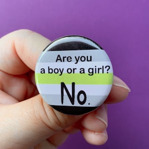 May include: A circular badge with a black rim, displaying the text "Are you a boy or a girl? No." The design incorporates horizontal stripes in white, grey, and a vibrant green.