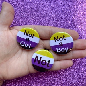 May include: Three round buttons with the text "Not a Girl", "Not a Boy", and "Not" on them. The buttons are yellow, white, purple, and black, and are arranged in a hand.