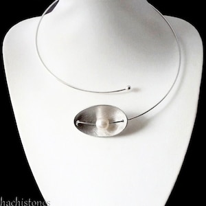 Könnte beinhalten: Eine silberfarbene Halskette mit minimalistischem Design. Die Halskette hat ein dünnes, gebogenes Band, das den Hals umschließt, mit einer kleinen Kugel auf der Rückseite. Ein ovaler Anhänger aus gebürstetem Metall hält eine einzelne, runde Perle. Der Schmuck wird auf einer weißen Büste präsentiert.