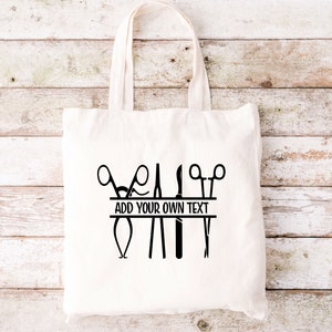 May include: White canvas tote bag with a black design of surgical instruments, including scissors, forceps, and a scalpel. The design includes a banner with the text "ADD YOUR OWN TEXT".