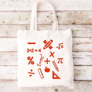 May include: White canvas tote bag with red mathematical symbols and equations, including a plus sign, minus sign, multiplication sign, division sign, percentage sign, pi symbol, square root symbol, and the equation E=mc2. The bag also features a red pencil, ruler, and apple.