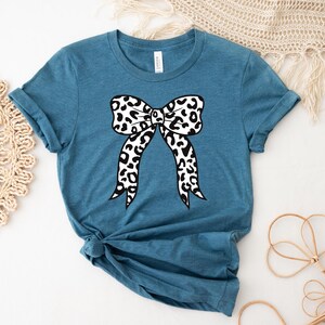 May include: A teal-colored t-shirt featuring a large bow design. The bow has a black and white animal print pattern. The shirt has short sleeves and a crew neck. The bottom of the shirt is tied in a knot.