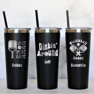 May include: Three black stainless steel tumblers with lids and straws. The tumblers are personalized with pickleball-themed designs and names. The first tumbler says "Just One More Game" with a pickleball paddle and ball. The second tumbler says "Dinkin' Around" with a pickleball ball. The third tumbler says "Pickleball Queen" with two crossed pickleball paddles and a ball.