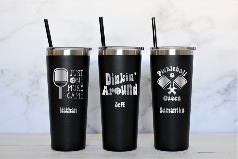 May include: Three black stainless steel tumblers with lids and straws. The tumblers are personalized with pickleball-themed designs and names. The first tumbler says "Just One More Game" with a pickleball paddle and ball. The second tumbler says "Dinkin' Around" with a pickleball ball. The third tumbler says "Pickleball Queen" with two crossed pickleball paddles and a ball.