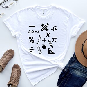 May include: White t-shirt with a black graphic design of math symbols and school supplies, including a pencil, ruler, apple, plus sign, minus sign, multiplication sign, division sign, percentage sign, pi symbol, square root symbol, and the equation E=mc&sup2;.