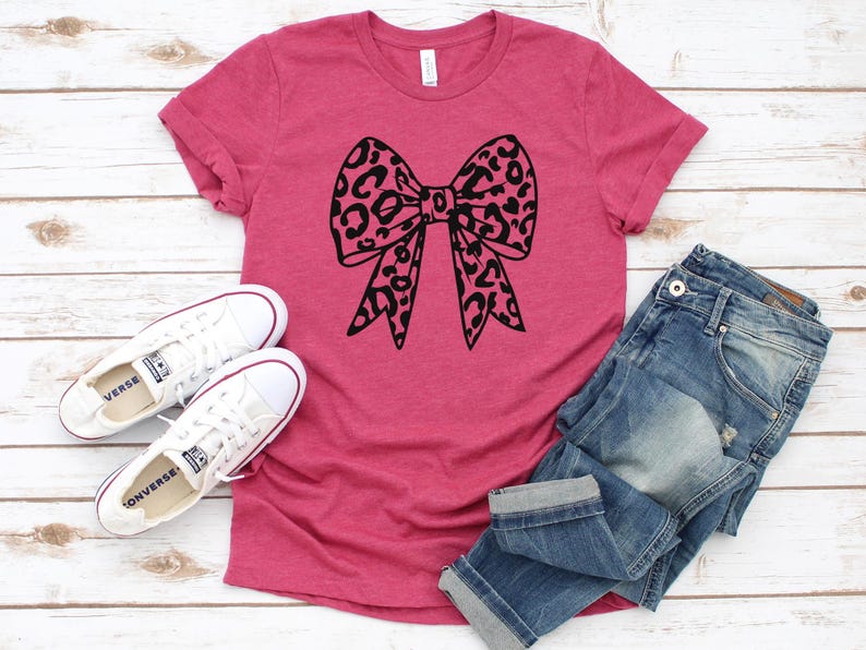 Pu&ograve; includere: Una maglietta rosa con un fiocco nero con stampa leopardata. La maglietta &egrave; appoggiata su una superficie di legno bianco con un paio di scarpe di tela bianche e un paio di jeans blu.