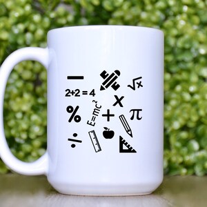 May include: White ceramic coffee mug with black mathematical symbols and icons, including a pencil, ruler, apple, plus sign, percentage sign, division sign, square root symbol, pi symbol, and the equation E=mc2.