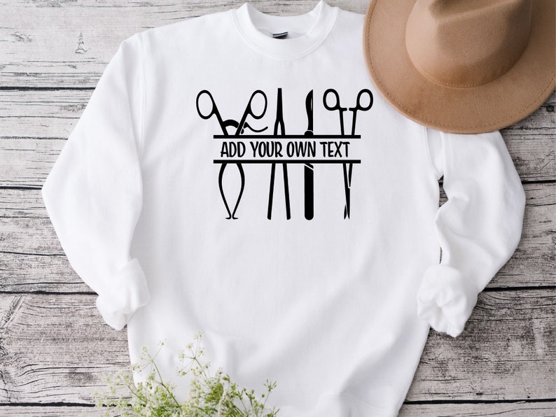 May include: White sweatshirt with a black design of surgical instruments, including scissors, forceps, and a scalpel. The design includes a banner that says "ADD YOUR OWN TEXT".