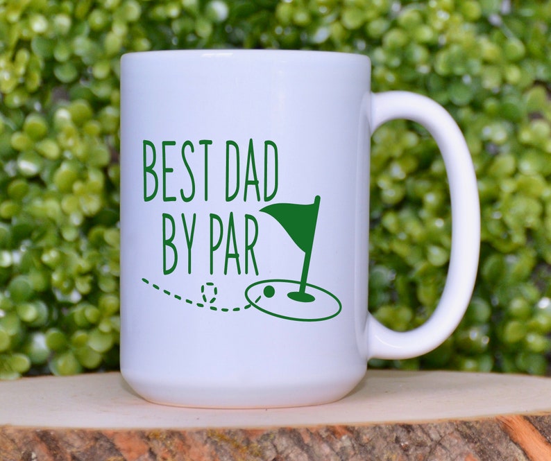 May include: White ceramic coffee mug with a green design that says "Best Dad By Par" with a golf ball and flag.