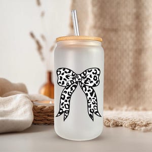 May include: Frosted glass can-shaped tumbler with a bamboo lid and a stainless steel straw. The tumbler features a black and white leopard print bow design. The background includes a neutral-toned fabric and decorative elements.