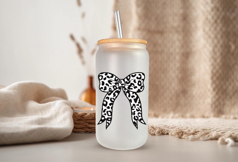 May include: Frosted glass can-shaped tumbler with a bamboo lid and a stainless steel straw. The tumbler features a black and white leopard print bow design. The background includes a neutral-toned fabric and decorative elements.