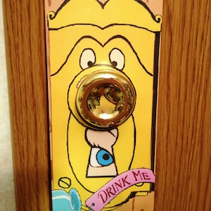 May include: A yellow door plate with a cartoon face design and a gold doorknob. The design includes a keyhole with a blue eye and the words "DRINK ME" on a pink tag. The plate is mounted on a wooden door.
