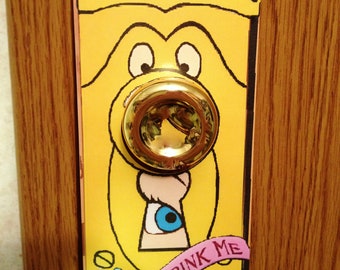 May include: A yellow door plate with a cartoon face design and a gold doorknob. The design includes a keyhole with a blue eye and the words "DRINK ME" on a pink tag. The plate is mounted on a wooden door.