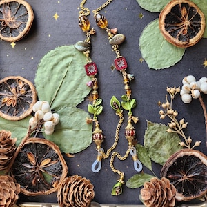May include: Gold-toned eyeglass chain with decorative beads and leaf accents. The chain features amber-colored and red beads, with green leaf charms. The chain is displayed on a dark surface with dried orange slices, leaves, and pinecones.