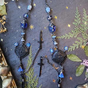 May include: Eyeglass chain with a dark silver chain, adorned with blue and clear beads, and decorative black charms. The chain features a small sword charm and is designed to hold eyeglasses.