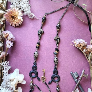 May include: A black metal eyeglass chain featuring black snake charms, ornate patterned beads, and small gold-toned skeleton key pendants. The chain is attached to a pair of thin-rimmed glasses, with the entire assembly resting on a purple surface.