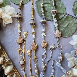 May include: Eyeglass chains with decorative beads, leaf charms, and small animal pendants. The chains are gold and silver, with clear rubber loops for attaching to glasses. The background includes dried leaves and a decorative frame.