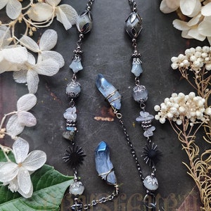 May include: A black chain with blue and white crystal beads and charms, including two large blue crystals, two black star charms, and two silver-toned clasps. The chain is laying on a dark surface with white flowers.