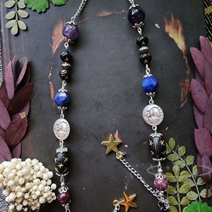 May include: Eyeglass chain with a celestial theme. The chain features a silver-tone metal chain with beads in shades of purple, black, blue, and silver. Gold star charms dangle from the chain, adding a touch of whimsy.