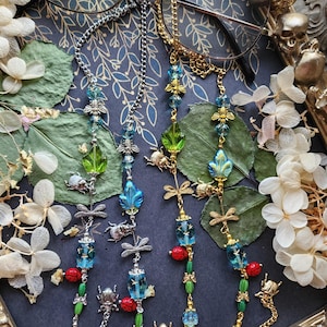 May include: Eyeglass chains with decorative insect and bead accents. The chains feature blue, green, and red beads, along with dragonfly, beetle, and bee charms. The chains are silver and gold in color, and are displayed on a decorative tray.