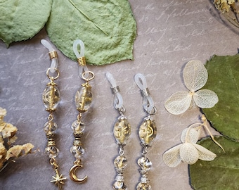 May include: Eyeglass chains with clear and gold beads, star and crescent moon charms. The chains have clear rubber loops to attach to glasses. The chains are displayed on a gray surface with dried flowers and leaves.