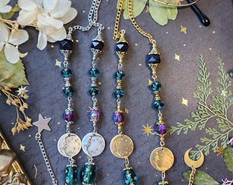 May include: Eyeglass chains with celestial-themed beads and charms. The chains feature a mix of silver and gold-toned metal, with beads in shades of blue, purple, and green. Moon and star charms add to the mystical aesthetic. The glasses are displayed on a dark background.