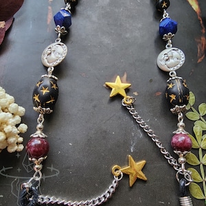 May include: A celestial-themed eyeglass chain featuring a mix of beads, including black, blue, and burgundy. The chain includes silver moon and star charms, with gold star accents. The chain is designed to hold eyeglasses.