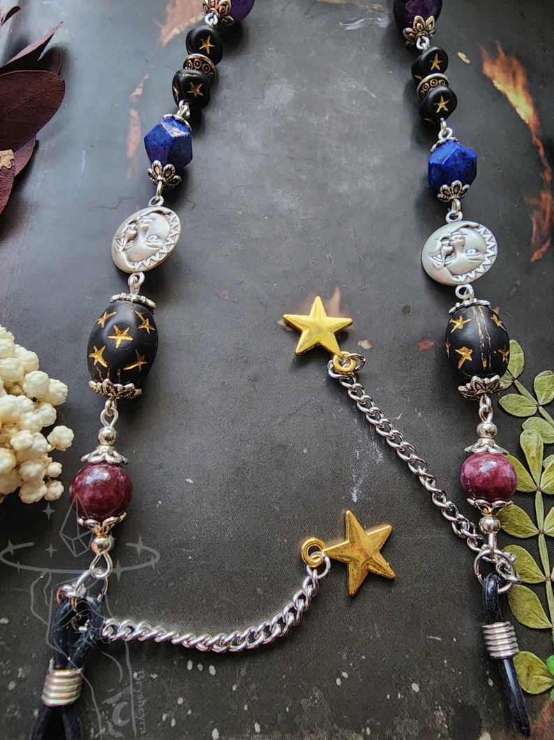May include: A celestial-themed eyeglass chain featuring a mix of beads, including black, blue, and burgundy. The chain includes silver moon and star charms, with gold star accents. The chain is designed to hold eyeglasses.