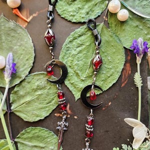 May include: A pair of eyeglass chains with black cords, featuring silver-toned metal charms. Each chain has a crescent moon, a red teardrop, and a dagger charm, accented with red beads. The chains rest on a dark surface with green leaves and white flowers.