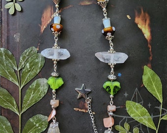 May include: A silver chain eyeglass holder with decorative beads and charms. The design includes clear quartz crystals, green leaf-shaped beads, and a star charm. The chain ends with a crescent moon charm and loops for glasses.