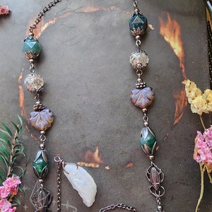 May include: Eyeglass chain featuring a dark metal chain, decorated with green, clear, and purple beads, and two iridescent crystal pendants. The chain has loops to attach to glasses and floral accents.