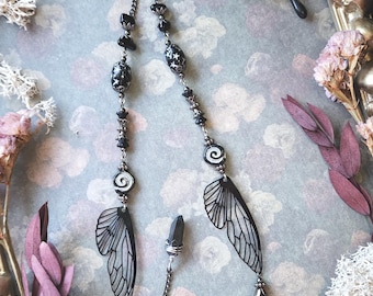 May include: A black metal eyeglass chain featuring two translucent black insect wing charms, two black and white spiral beads, two black star-patterned beads, and two dark crystal points wrapped in silver wire, finished with black rubber loops.