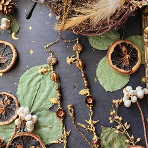 May include: A gold-coloured glasses chain featuring leaf and bird charms, and brown beads. The chain is displayed on a dark surface with dried citrus slices, pine cones, and greenery.