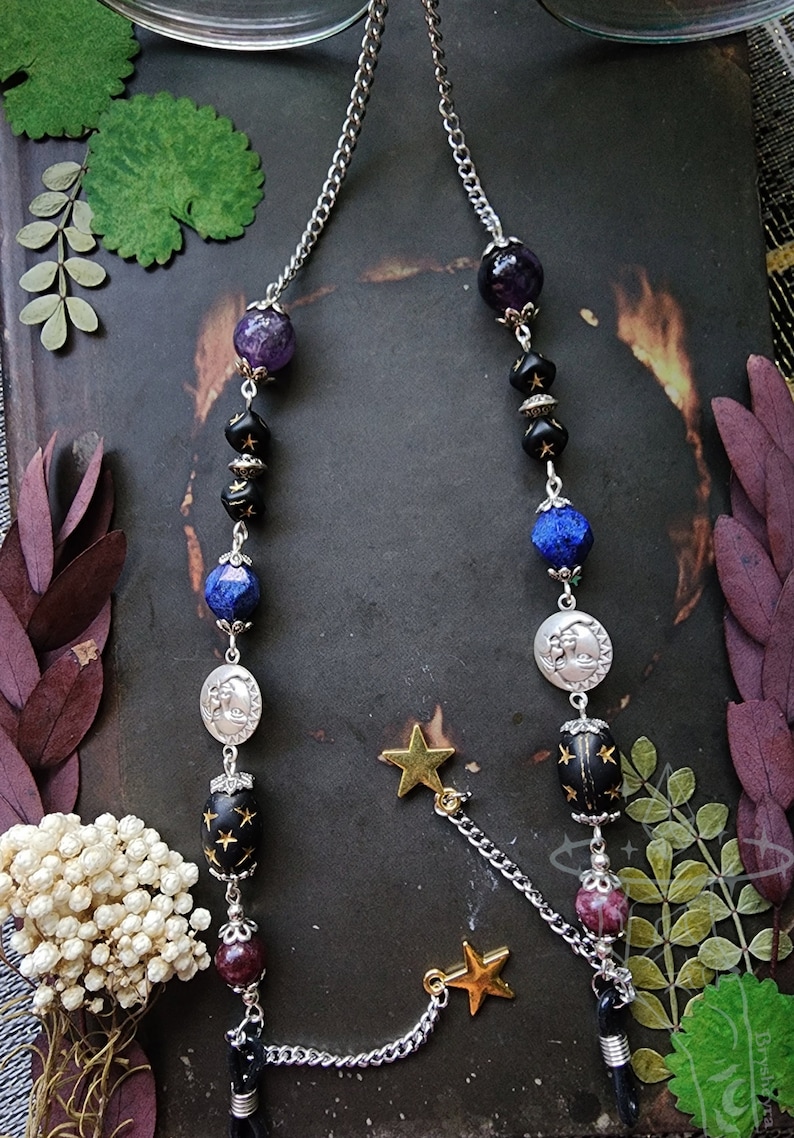 May include: Eyeglass chain with a celestial theme. The chain features a silver-tone metal chain with beads in shades of purple, black, blue, and silver. Gold star charms dangle from the chain, adding a touch of whimsy.
