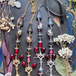 May include: Eyeglass chains with decorative pendants. The chains feature gold and silver-toned metal, with black, red, and iridescent beads. Some pendants include skull and leaf designs. The glasses are resting on a dark, textured surface.