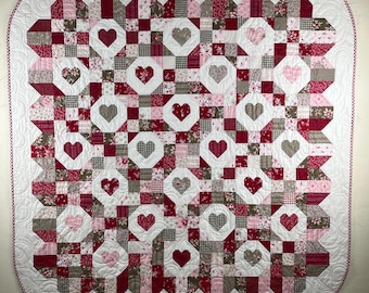 May include: A handmade quilt featuring a repeating heart pattern. The quilt showcases a patchwork design with red, pink, white, and gray fabrics. The hearts are in various shades, and the quilt has a white border with a red trim.