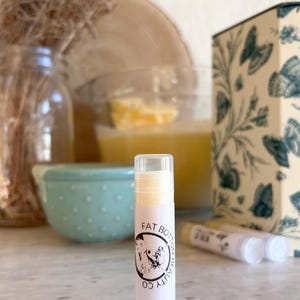 May include: A white tube of lip balm with a black and white label that says "Fat Bottom Beauty Co.". The lip balm is sitting on a white surface.