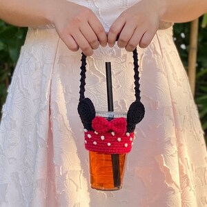 May include: A crocheted Minnie Mouse cup holder with black ears, a red bow, and white polka dots. The holder is attached to a black strap and is holding a clear plastic cup with a straw and a brown beverage.